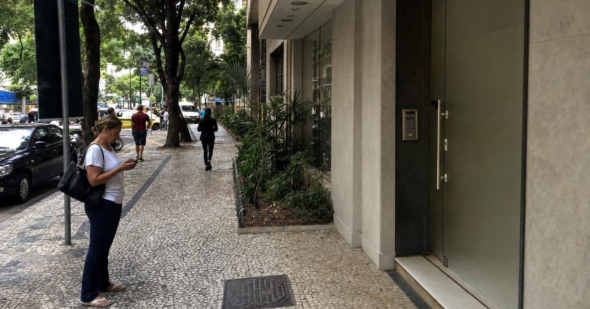 Calçada em frente a um prédio de consultórios no Rio de Janeiro, com uma mulher no ponto de ônibus pesquisando no celular.