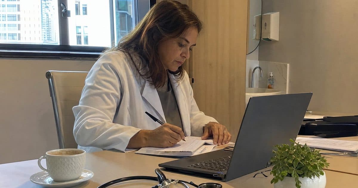 Médica sozinha no consultório no fim do dia, revendo anotações num caderno ao lado do notebook.